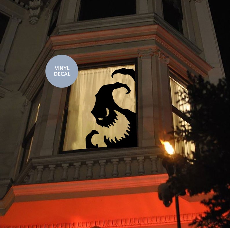 Op de afbeelding: Een zwarte vinyl sticker met het silhouet van een griezelig monster, geplaatst op een raam van een huis. De sticker wordt van achteren verlicht, waardoor een griezelig effect ontstaat.