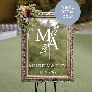 May include: White vinyl decal with the letters "M" and "A" intertwined with a leafy branch design. The text "Maureen & Alex" and the date "11.20.20" are below the initials. The decal is on a clear glass mirror with a gold ornate frame.