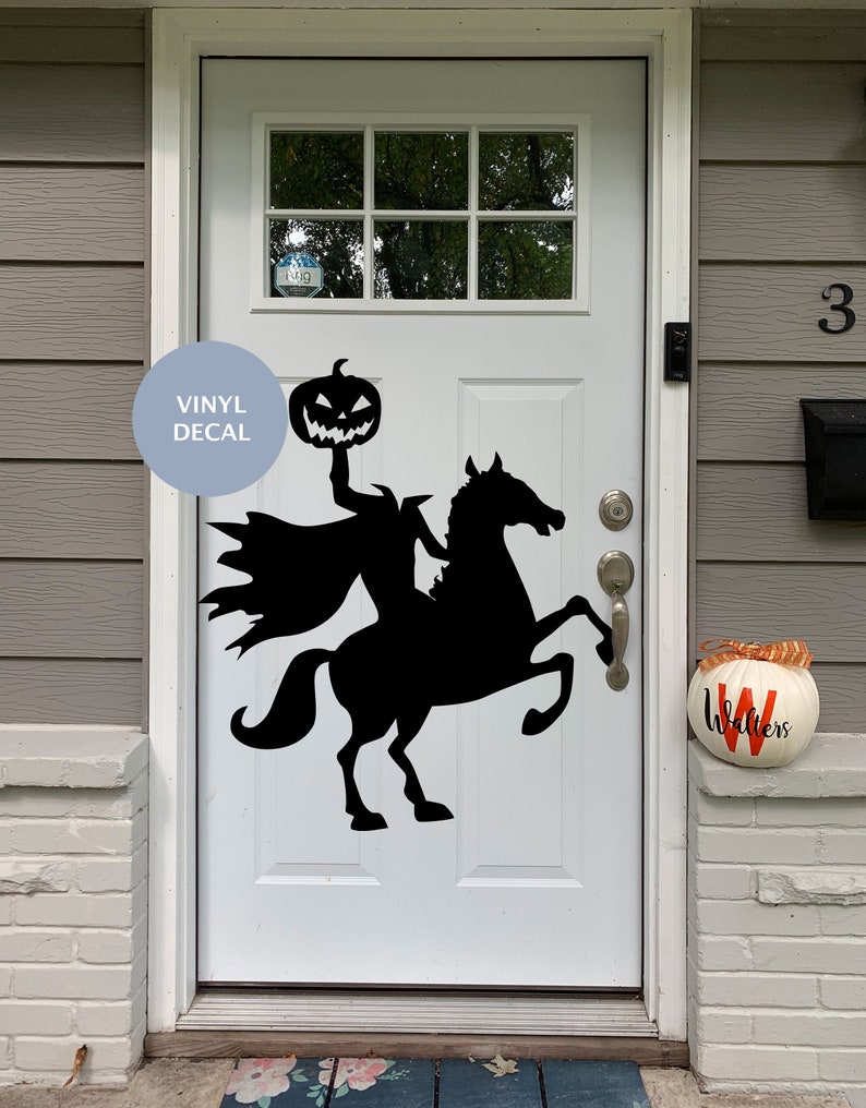 Puede incluir: Un vinilo adhesivo negro aplicado sobre una puerta blanca, que representa a un jinete sin cabeza montando un caballo mientras sostiene una calabaza tallada. El dise&ntilde;o muestra una capa ondeante y la silueta de un caballo encabritado, creando un contraste gr&aacute;fico oscuro sobre la superficie blanca.