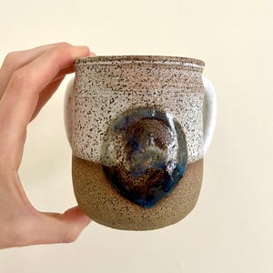May include: A handmade ceramic mug with a speckled white and brown exterior. The bottom half is a solid brown, and a dark blue glaze detail is on the front. The mug has two white handles.