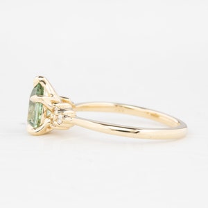May include: A gold ring featuring a pear-shaped, light green gemstone as the centerpiece. The ring has a delicate band and small, round accent stones on either side of the main stone. The setting is intricate and detailed.