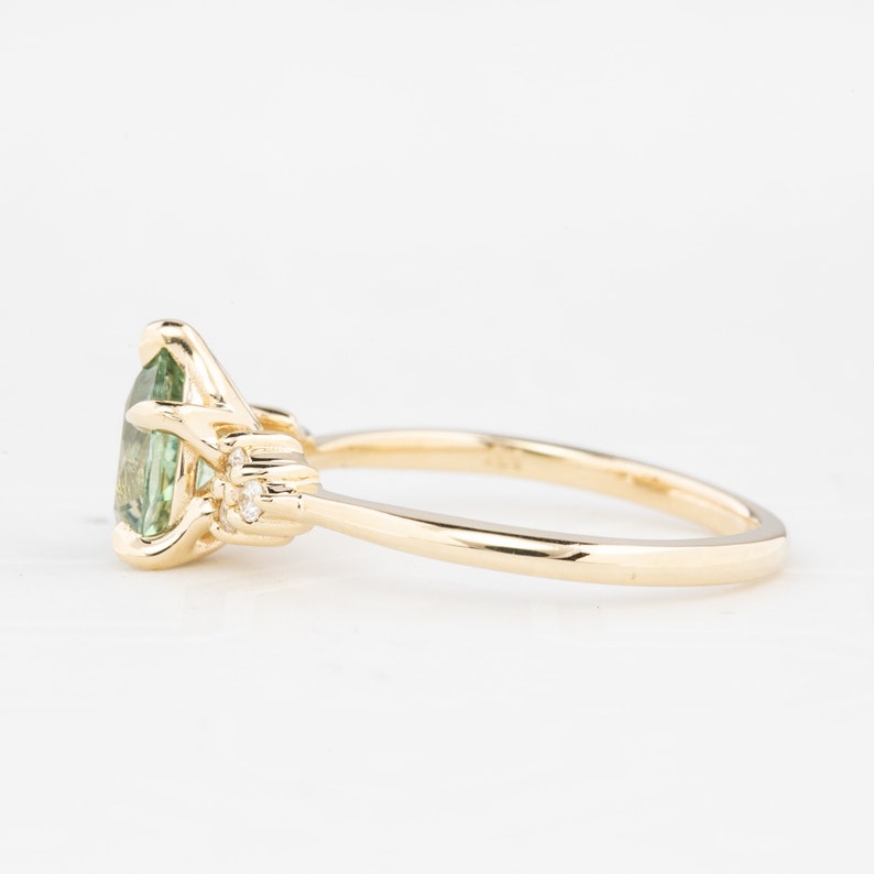 May include: A gold ring featuring a pear-shaped, light green gemstone as the centerpiece. The ring has a delicate band and small, round accent stones on either side of the main stone. The setting is intricate and detailed.