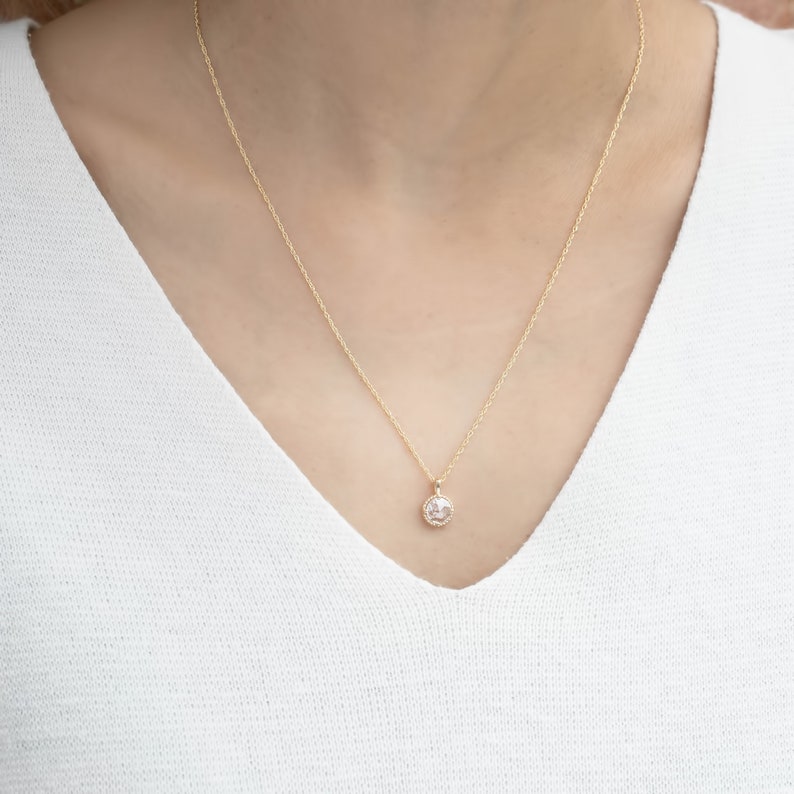 May include: A delicate gold chain necklace with a small round pendant featuring a clear crystal surrounded by a halo of smaller crystals.