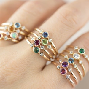 May include: Five gold rings with a delicate design featuring a cluster of small diamonds and a single larger gemstone in each ring. The gemstones are blue, green, red, purple, and orange.