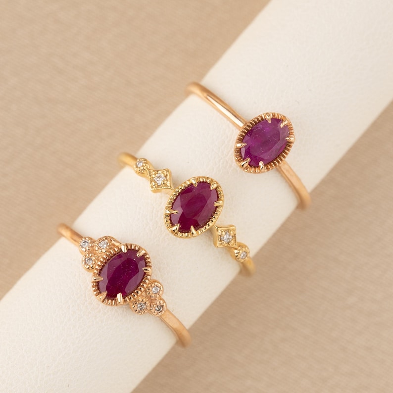 May include: Three gold rings with oval-shaped ruby gemstones and diamond accents. The rings are displayed on a white surface.