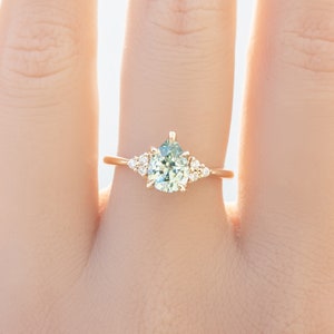 May include: A gold ring featuring a pear-shaped, light green gemstone as the centerpiece, flanked by smaller, round, clear gemstones. The ring's band is a delicate gold color, and the design is set against a soft, neutral background.