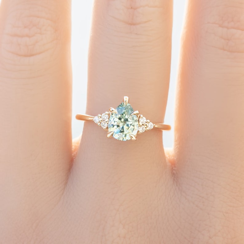 May include: A gold ring featuring a pear-shaped, light green gemstone as the centerpiece, flanked by smaller, round, clear gemstones. The ring's band is a delicate gold color, and the design is set against a soft, neutral background.
