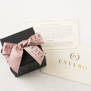 May include: A black gift box with a pink ribbon tied around it. The box has the text "Envero New York" printed on it. The box is sitting on a white surface next to a white card with the text "Envero New York" printed on it.