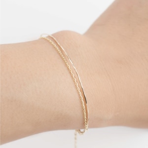May include: Two gold bracelets on a person's wrist. The bracelets are layered, one is a delicate chain and the other is a simple, thin band.