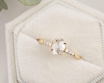 May include: A gold ring featuring a large, oval-cut, clear gemstone. The band is embellished with small, round gemstones. The ring is displayed in a white velvet jewellery box, with a sprig of greenery in the background.