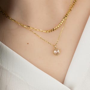 May include: A gold chain necklace with a delicate pendant featuring a clear round gemstone surrounded by a gold halo.