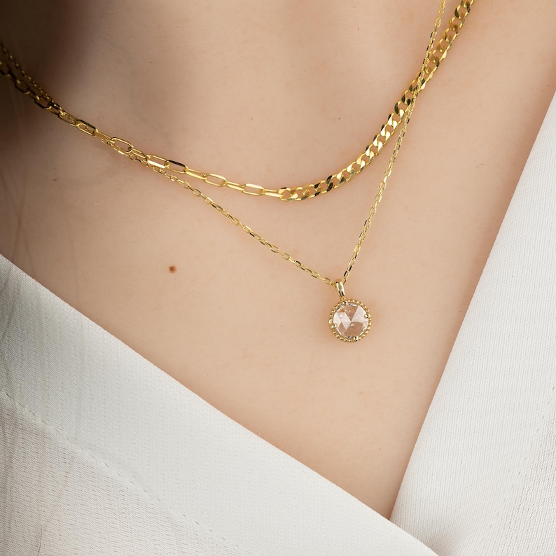 May include: A gold chain necklace with a delicate pendant featuring a clear round gemstone surrounded by a gold halo.