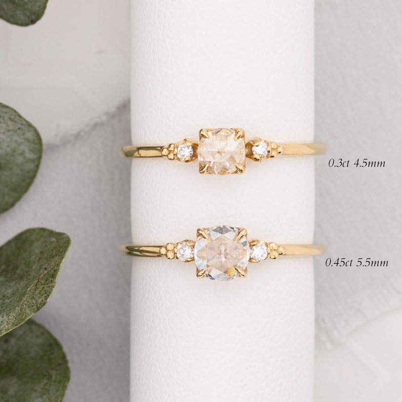 May include: Two gold rings with a single round diamond in the center and two smaller diamonds on either side. The top ring has a 0.3ct 4.5mm diamond and the bottom ring has a 0.45ct 5.5mm diamond.