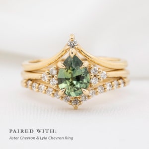 May include: A set of three gold rings. The engagement ring features a pear-shaped green gemstone surrounded by small diamonds. The other two rings have a chevron design. The text at the bottom reads "PAIRED WITH: Aster Chevron & Lyla Chevron Ring."