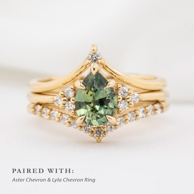 May include: A set of three gold rings. The engagement ring features a pear-shaped green gemstone surrounded by small diamonds. The other two rings have a chevron design. The text at the bottom reads "PAIRED WITH: Aster Chevron & Lyla Chevron Ring."