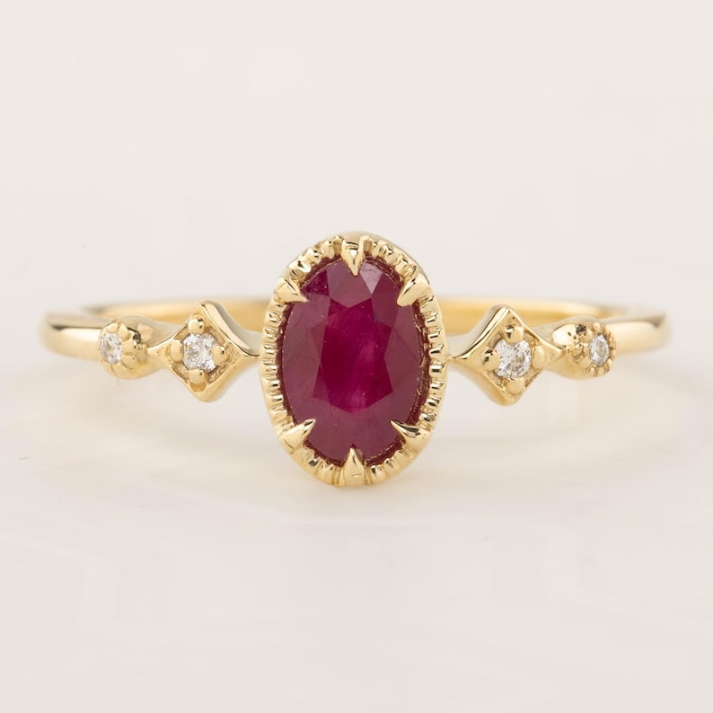 May include: A gold ring with a red oval ruby gemstone surrounded by a delicate gold setting. The ring has a band with small diamond accents.