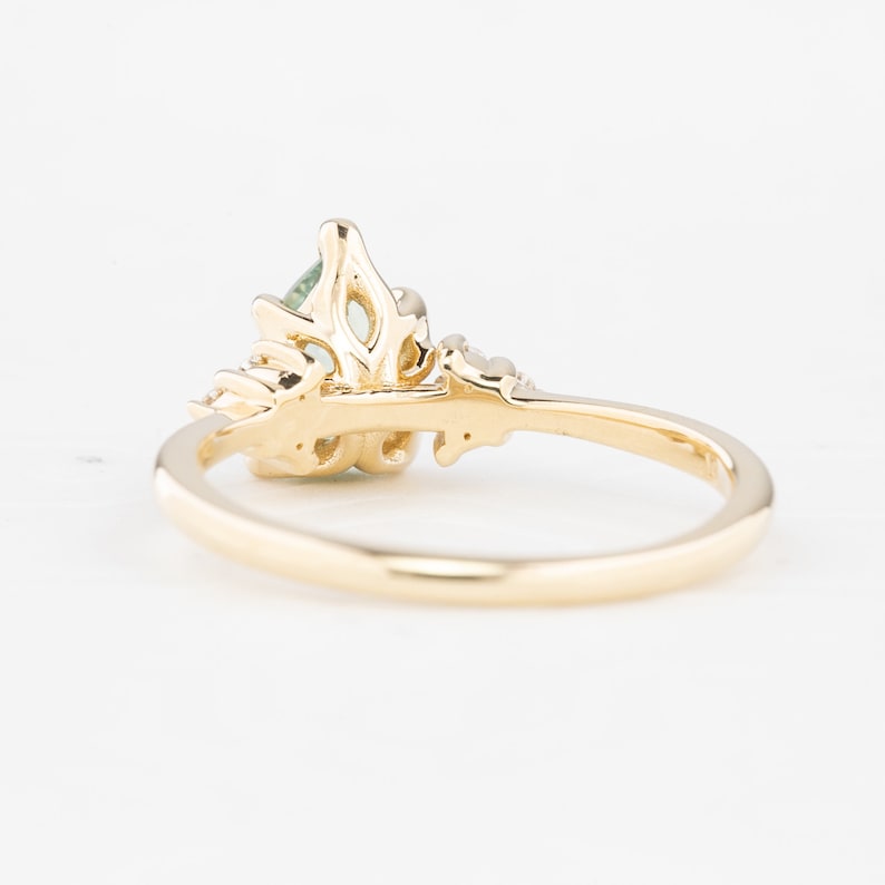 May include: A gold ring with a pear-shaped, light green gemstone. The ring features intricate detailing around the gemstone and along the band, with small accent stones. The ring is set against a white background.