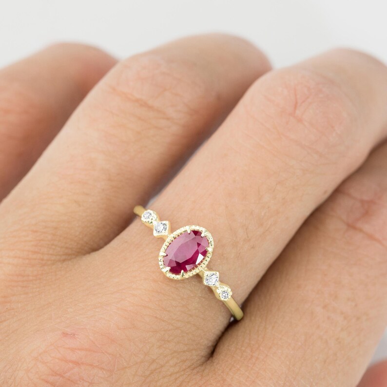 May include: A gold ring with an oval ruby gemstone surrounded by small diamonds. The ring is set on a finger.