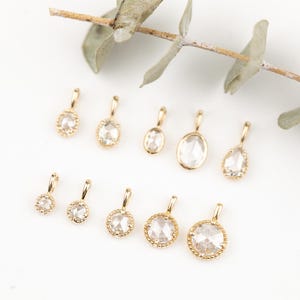May include: A collection of gold pendants with clear, faceted gemstones in various shapes, including round, oval, and teardrop. Each pendant has a small loop for a chain. The pendants are displayed on a white surface with a sprig of greenery.