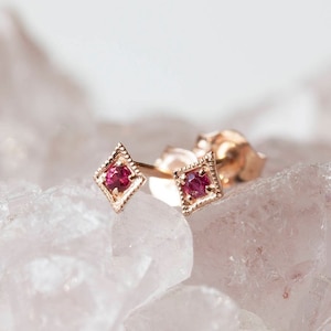 May include: A pair of small, delicate earrings with a square-shaped setting and a single red gemstone in each. The earrings are made of gold and have a simple, elegant design.