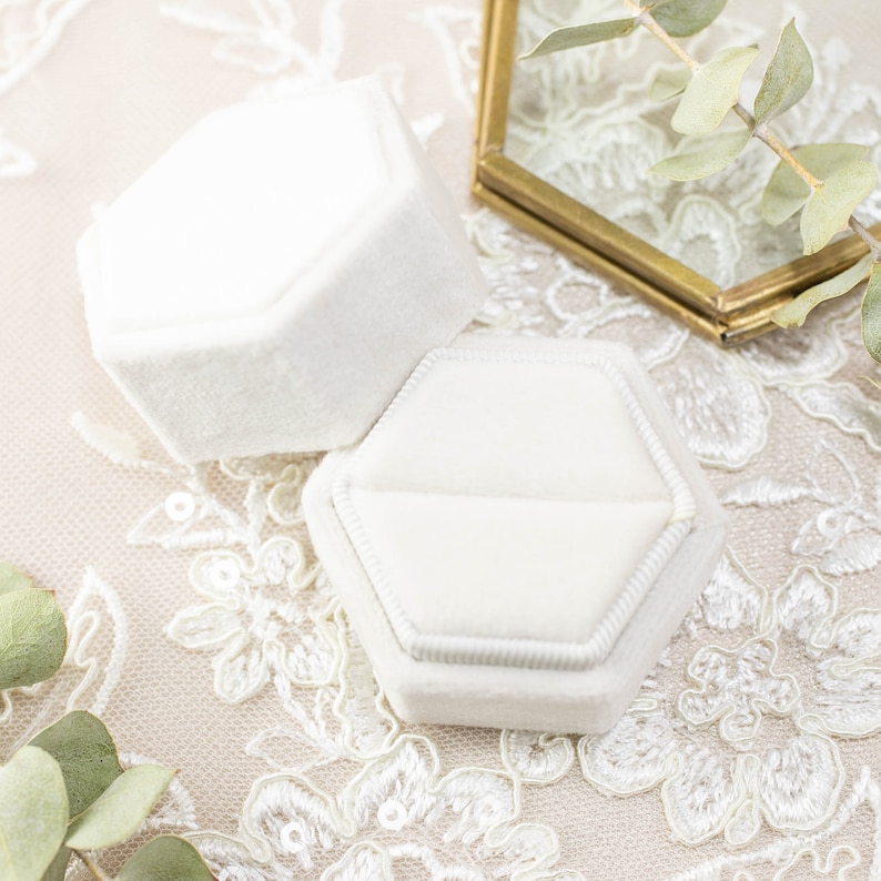 May include: Two white velvet hexagonal ring boxes. One box is open, revealing the interior. The boxes are on a white lace surface, with a gold-framed glass geometric shape and green leaves in the background.