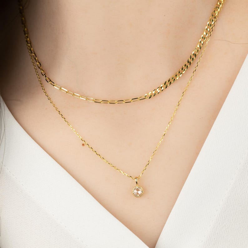 May include: A gold chain necklace with a small, round pendant featuring a clear gemstone. The necklace is layered over a thicker gold chain.
