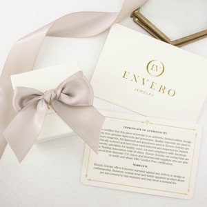 May include: A white box with a beige ribbon tied in a bow. The box is sitting on a white surface. A white card with gold lettering reads "Envero Jewelry" and "Certificate of Authenticity". The card includes a 6-month warranty against defects in design or craftsmanship.