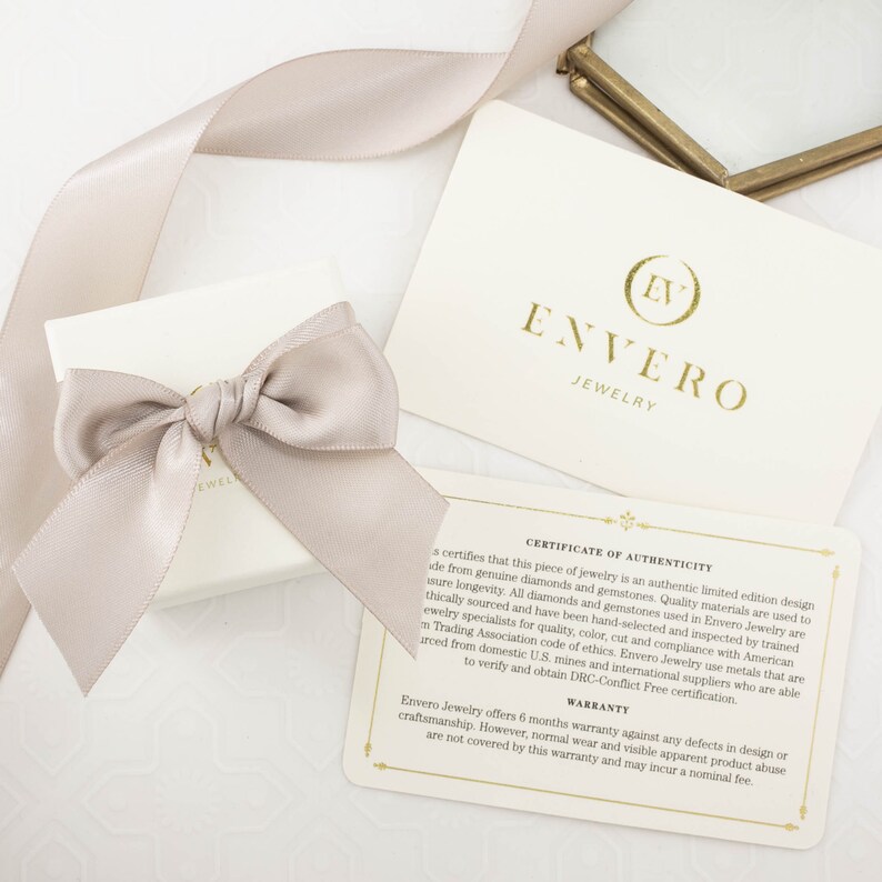 May include: A white box with a beige ribbon tied in a bow. The box is sitting on a white surface. A white card with gold lettering reads "Envero Jewelry" and "Certificate of Authenticity". The card includes a 6-month warranty against defects in design or craftsmanship.