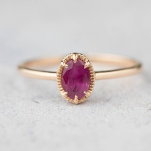 May include: A delicate gold ring with a single oval-shaped ruby gemstone set in a detailed, ornate setting.