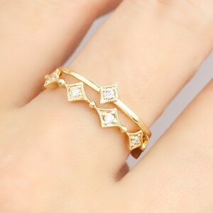 May include: A gold ring with a delicate design featuring small diamonds set in a square shape. The ring is worn on a finger.