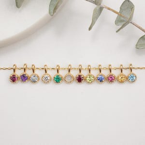May include: A gold chain with 10 small charms, each featuring a different colored gemstone. The gemstones are set in a gold bezel and have a scalloped edge. The charms are arranged in a row on the chain.