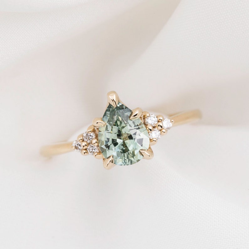 May include: A gold ring featuring a pear-shaped, light green gemstone as the centerpiece, accented with small, round, clear gemstones. The ring has a delicate band and is set against a white background.