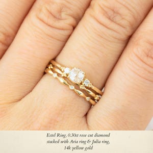 May include: A gold ring set with a rose cut diamond, stacked with two smaller diamond rings. The rings are on a finger and the text "Estel Ring, 0.30ct rose cut diamond stacked with Aria ring & Julia ring, 14k yellow gold" is below the image.