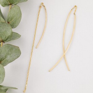 May include: A pair of delicate gold-colored earrings. One earring features a chain, while the other has a curved, elongated design. The earrings are displayed against a white background, with a sprig of green leaves in the upper left corner.