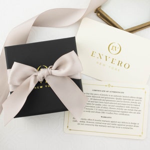 May include: A black jewelry box with a beige satin ribbon tied in a bow. The box is labeled "Envero New York". The box is sitting on a white surface next to a card that reads "Certificate of Authenticity" and "Warranty".