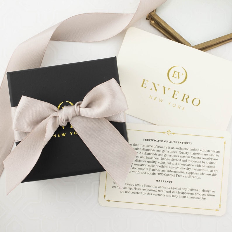 May include: A black jewelry box with a beige satin ribbon tied in a bow. The box is labeled "Envero New York". The box is sitting on a white surface next to a card that reads "Certificate of Authenticity" and "Warranty".
