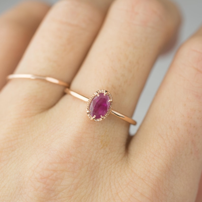 May include: A delicate rose gold ring with a vibrant oval ruby gemstone set in a delicate, textured bezel setting.
