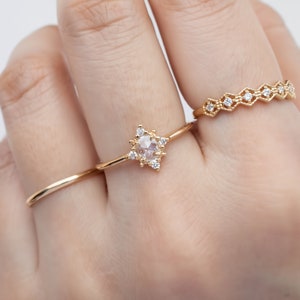 May include: A gold ring with a star-shaped setting featuring a round, clear gemstone surrounded by smaller clear gemstones. The ring is worn on a finger with two other gold rings.