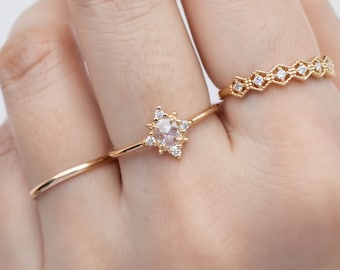 May include: A gold ring with a star-shaped setting featuring a round, clear gemstone surrounded by smaller clear gemstones. The ring is worn on a finger with two other gold rings.