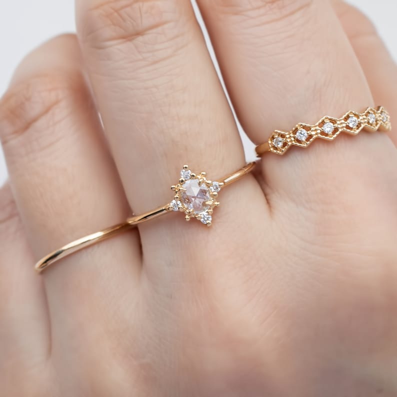 May include: A gold ring with a star-shaped setting featuring a round, clear gemstone surrounded by smaller clear gemstones. The ring is worn on a finger with two other gold rings.