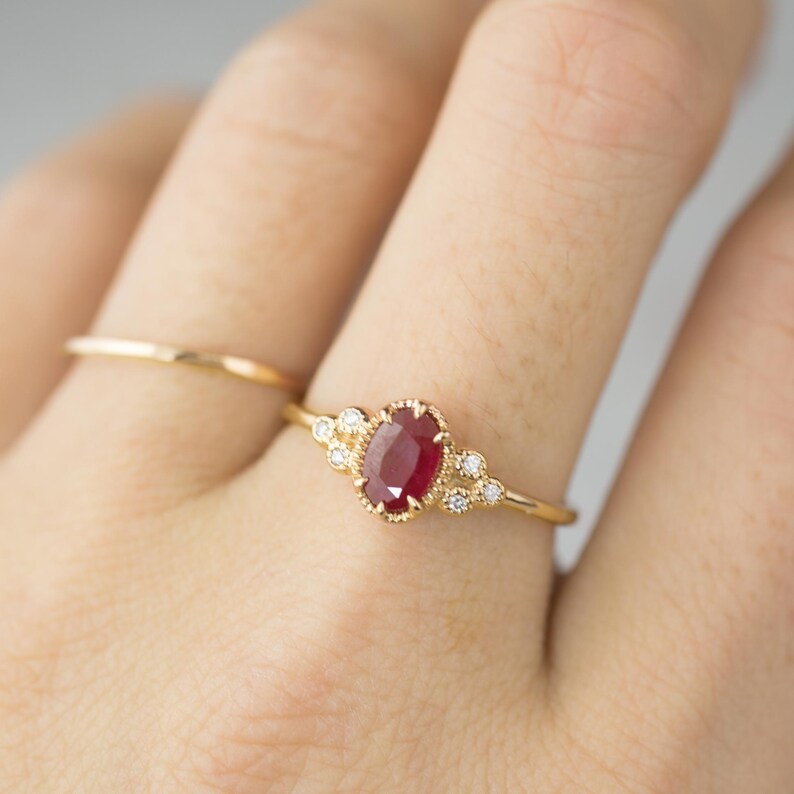May include: A gold ring with a red oval ruby gemstone surrounded by smaller diamonds. The ring is on a finger.
