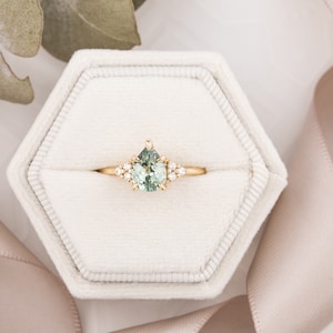 May include: A gold ring with a pear-shaped, light green gemstone as the focal point. The ring is set with small, round, clear gemstones on either side of the main stone. The ring is displayed in a white, hexagonal ring box.