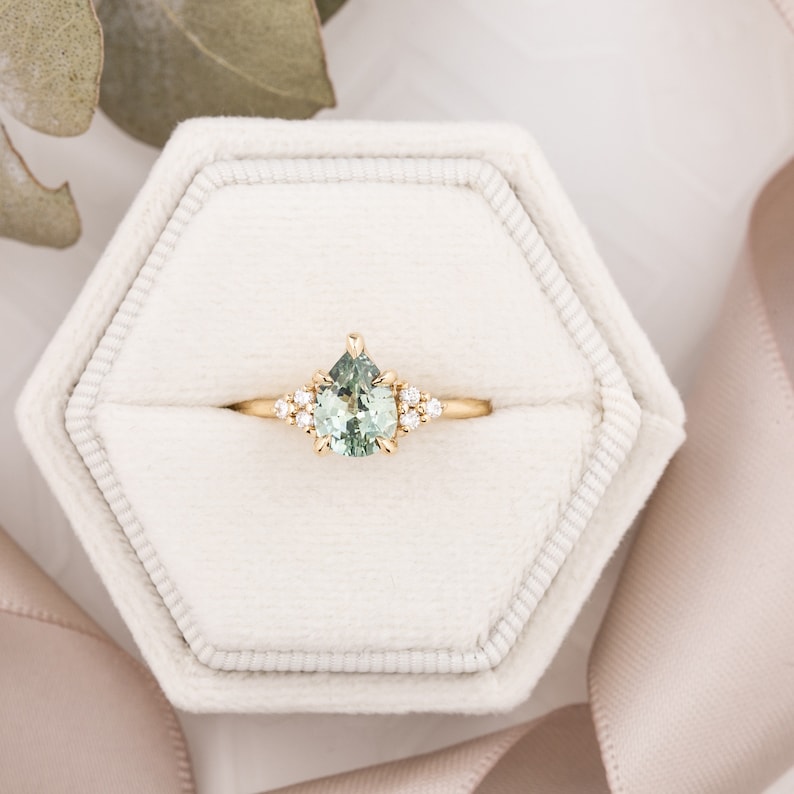 May include: A gold ring with a pear-shaped, light green gemstone as the focal point. The ring is set with small, round, clear gemstones on either side of the main stone. The ring is displayed in a white, hexagonal ring box.
