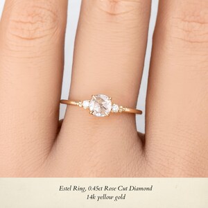 May include: A delicate gold ring with a rose cut diamond and two smaller accent diamonds. The ring is on a person's finger.