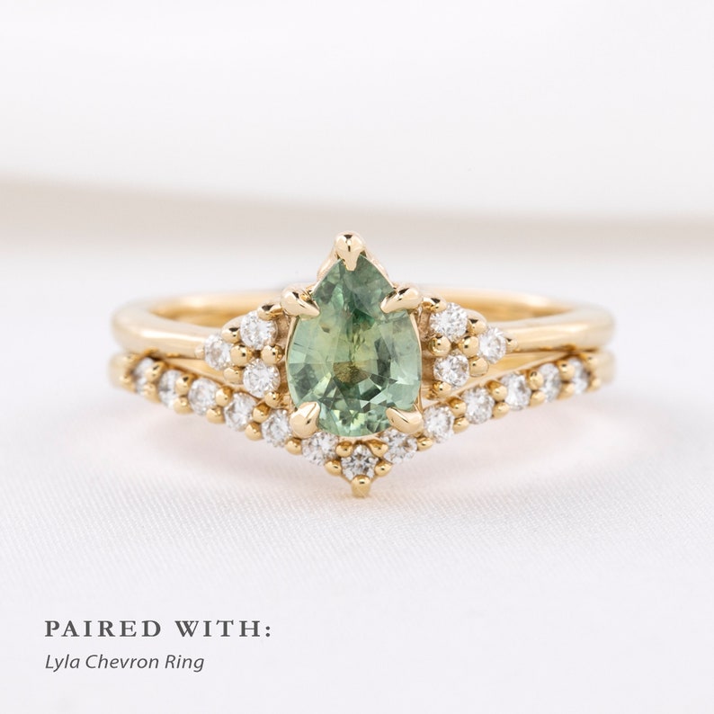May include: A gold engagement ring with a pear-shaped green gemstone, surrounded by small white diamonds. The ring is paired with a gold chevron ring, also adorned with small white diamonds. Text on the image reads "PAIRED WITH: Lyla Chevron Ring."