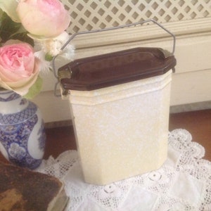May include: A white ceramic container with a brown lid and a metal handle. The container has a speckled pattern and is sitting on a white lace doily.