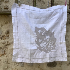 May include: White linen square with a floral design in the center. The edges are finished with a delicate stitched border. The cloth is hanging on a wooden structure with a stone wall in the background.