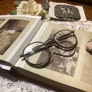May include: An open antique photo album with sepia-toned images, featuring a pair of round, wire-rimmed spectacles. A decorative metal buckle and a cream-colored rose are also visible, suggesting a vintage theme.