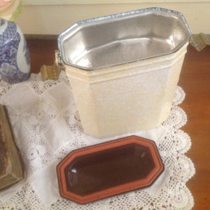 May include: A white metal container with a silver interior and a brown ceramic dish. The container has a lid with a handle and a decorative design.