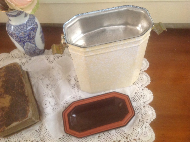 May include: A white metal container with a silver interior and a brown ceramic dish. The container has a lid with a handle and a decorative design.